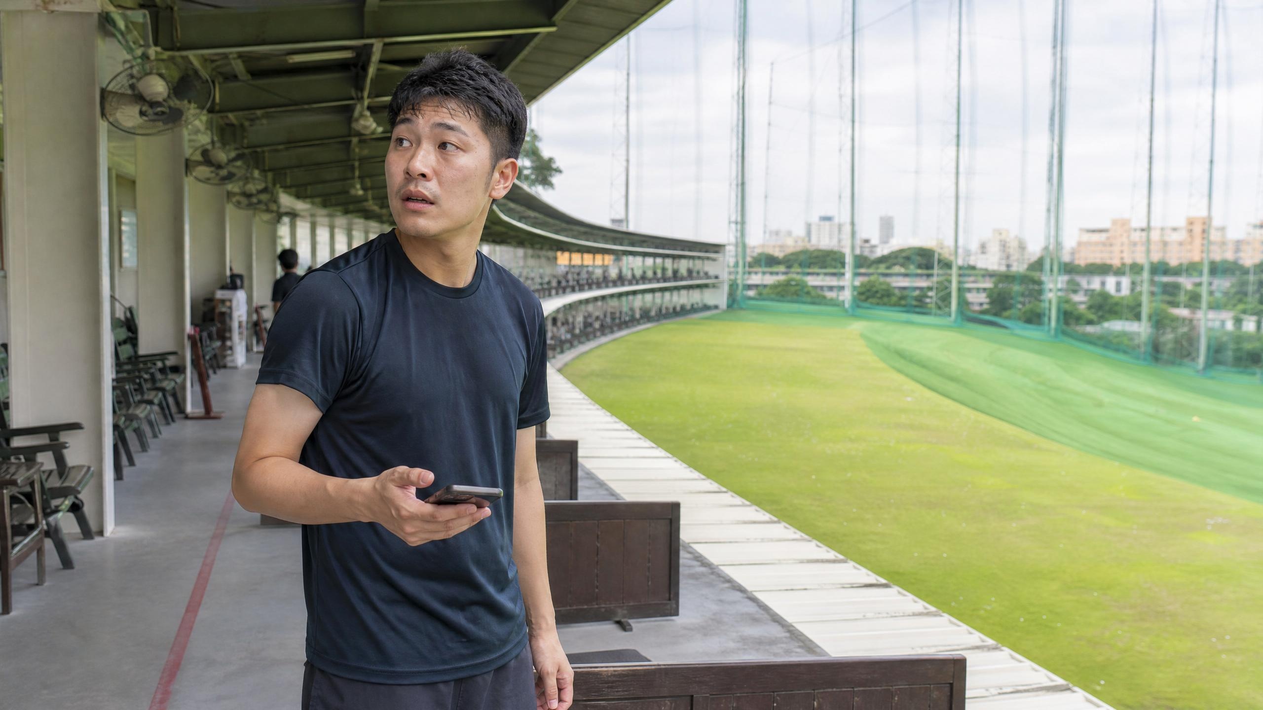 很多新手第一次來練習場就是這個狀態——不知道要問誰、怎麼開口。其實直接問櫃台就好。 一個台灣男生拿著手機站在練習場走道上,表情有點茫然不知道下一步怎麼辦