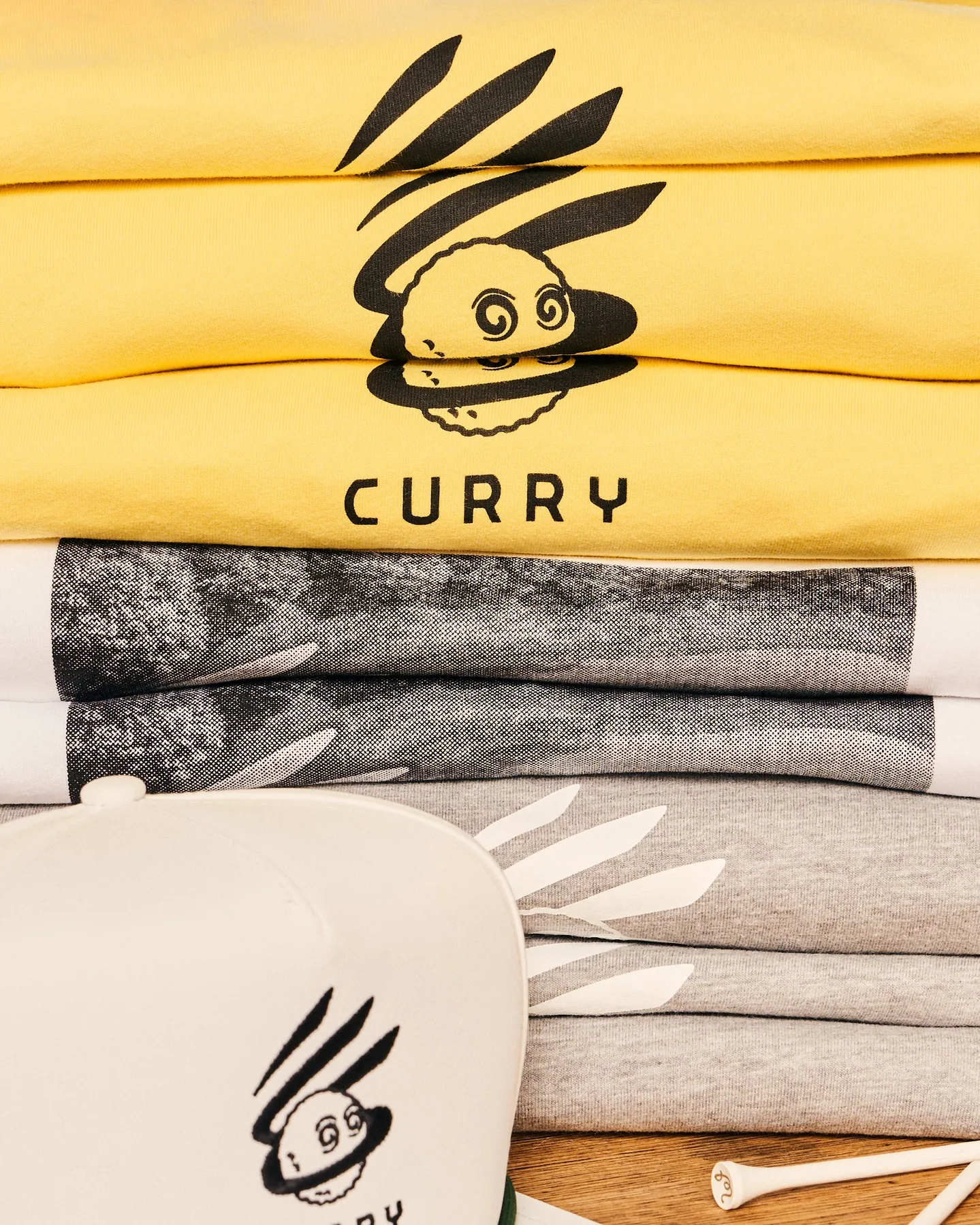 Curry Brand × Malbon 聯名黃色 T 恤與帽 T 疊放，印著 CURRY 小人圖案